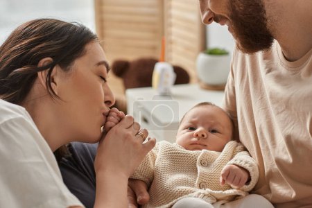 fröhliche Mutter küsst kleine Hand ihres Neugeborenen, während ihr Mann ihn hält, Familienkonzept