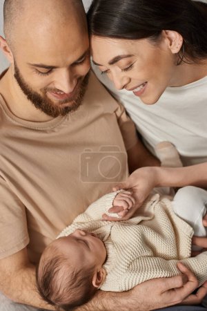 vertikale Aufnahme junger moderner Eltern, die ihr neugeborenes Baby halten und es liebevoll anlächeln, Familie