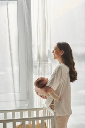 Photo for Joyous beautiful mother holding her newborn son next to his crib and smiling happily, family - Royalty Free Image