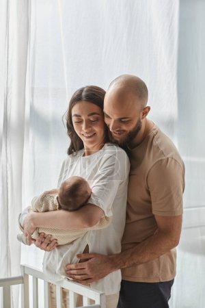 vertikale Aufnahme eines glücklichen Paares, das seinen neugeborenen Jungen hält und ihn neben der Krippe anlächelt, Familie