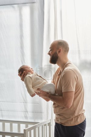 Vertikale Aufnahme eines glücklichen Vaters in kuscheliger Hauskleidung, der seinen neugeborenen Sohn hält und ihn liebevoll anlächelt