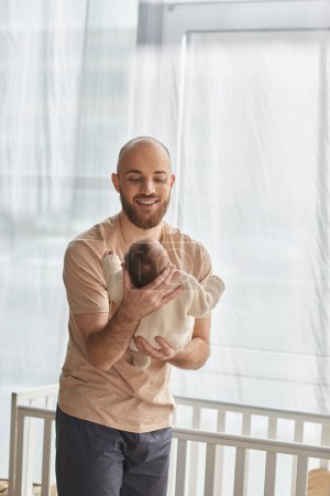 vertical shot of good looking bearded father holding his baby boy next to crib, family concept