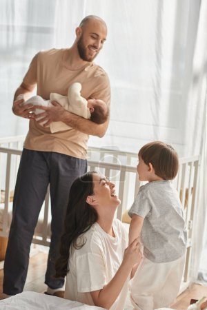 vertical shot of jolly father holding baby next to his beautiful wife playing with their little son