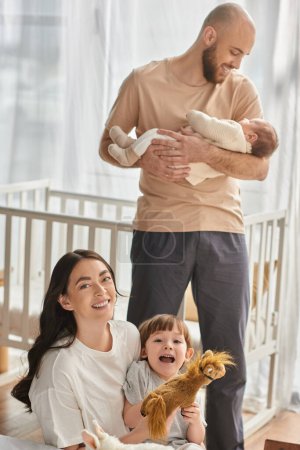 centrarse en la madre jugando con su hijo y sonriendo a la cámara junto a su marido borroso sosteniendo al bebé