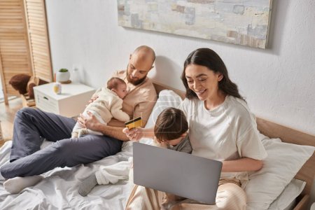 joyous relaxing family spending time together in bed with laptop and credit card, modern parenting