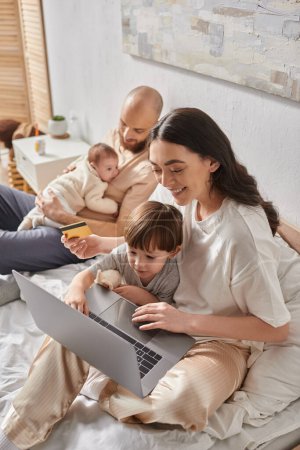 focus on mother with little son with laptop and credit card next to blurred husband and newborn