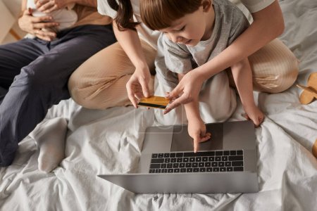 top view of little boy pointing at laptop surrounded by family with his mother holding credit card
