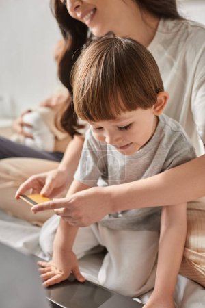 vertical focused shot of little boy looking at laptop next to his blurred mother holding credit card