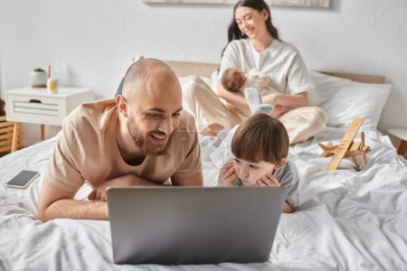 focus on jolly man watching movie on laptop with his son next to his blurred wife holding baby