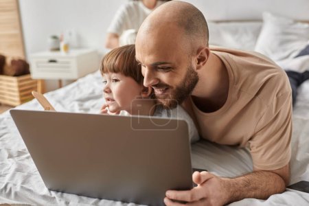 feliz padre barbudo abrazando a su pequeño hijo y mirando alegremente a la computadora portátil, concepto de familia