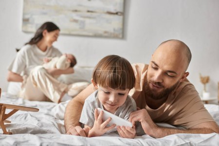 modern father looking at phone with his little son with his blurred wife and baby on backdrop