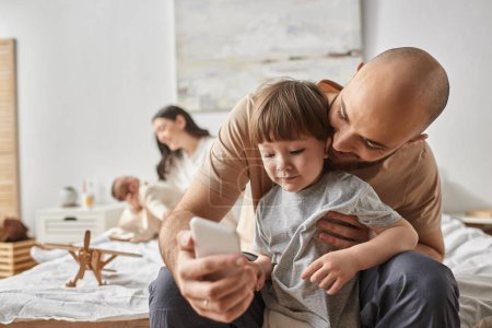 focus on jolly father and his little son looking at phone near his blurred wife and baby on backdrop