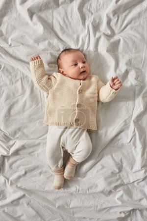 vista superior de lindo bebé recién nacido niño en cárdigan beige acostado sobre una manta blanca y mirando hacia otro lado
