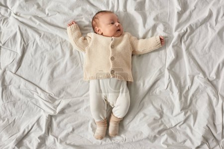 top view of adorable newborn baby boy in beige warm cardigan lying on white blanket with raised arms