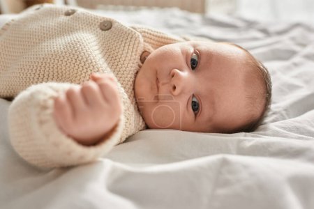 Porträt eines entzückenden kleinen neugeborenen Jungen in beiger Strickjacke, der auf dem Bett liegt und in die Kamera blickt