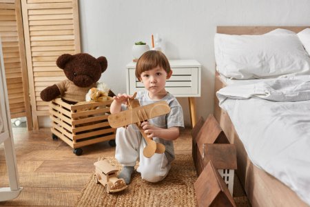 adorable niño preescolar en ropa de casa sosteniendo su juguete plano de madera y mirando directamente a la cámara