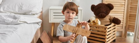 adorable niño en acogedora ropa de casa sosteniendo su juguete plano de madera y mirando a la cámara, pancarta