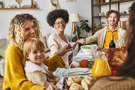joyeuse famille multiethnique assise tenant la main et priant à la table de Thanksgiving, reconnaissante pour le repas