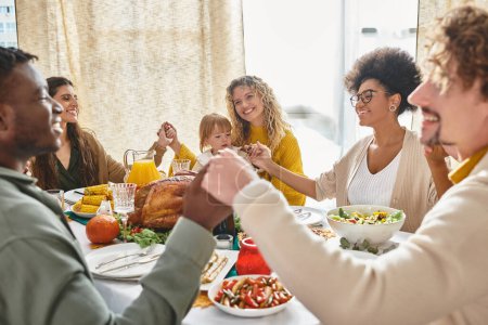 fröhliche multiethnische Freunde und Familie, die sich an den Händen halten und gemeinsam am Thanksgiving-Tisch beten