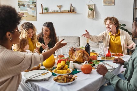 heureuse famille multiraciale ayant une conversation active et gestuelle à la table de Thanksgiving avec la dinde