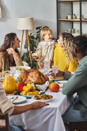 Celebración de Acción de Gracias, familia interracial feliz mirando a la niña cerca de la mesa festiva