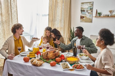 Téléchargez les photos : Homme heureux passant plaque avec pommes de terre rôties à un ami afro-américain pendant le dîner de Thanksgiving - en image libre de droit