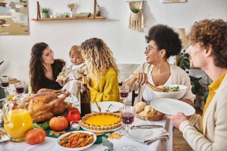 multicultural friends having delicious dinner while gathering on Thanksgiving, women calming child