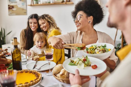 joyful african american woman serving salad to curly man on Thanksgiving day, lgbt family and baby