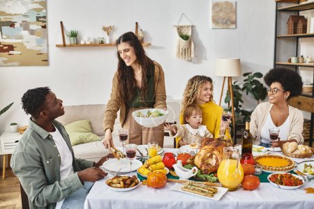 glückliche Frau serviert afrikanisch-amerikanischen Mann in der Nähe von Freunden und Familie am Thanksgiving-Tag Salat