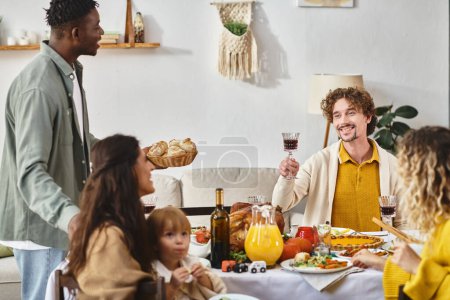 cheerful man toasting with glass of red wine near multicultural friends and family on Thanksgiving