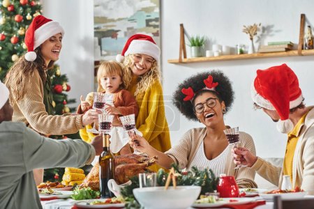 gran familia alegre multiétnica tintineando sus gafas en la mesa festiva con sombreros de Santa Claus, Navidad