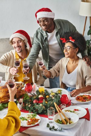 Téléchargez les photos : Parents multiethniques joyeux en tenue décontractée avec chapeaux de Père Noël accrochant leurs lunettes, Noël - en image libre de droit