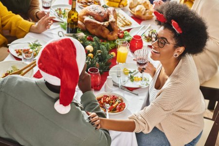 Foto de Alegre pareja afroamericana en la mesa de Navidad con su familia multiétnica, mano a hombro - Imagen libre de derechos