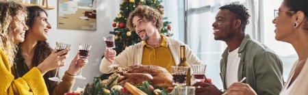 fröhliche multiethnische Verwandte beim Essen und Anstoßen mit Wein mit Weihnachtsbaum auf dem Hintergrund, Banner