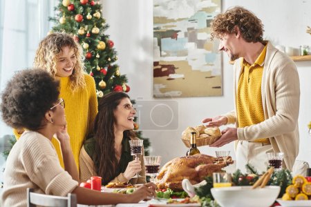 joven hombre alegre sosteniendo plato de pan rodeado de su familia multiétnica, árbol de Navidad telón de fondo