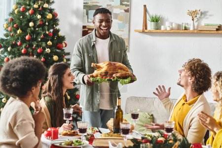 joyous african american surrounded by his multicultural family holding plate with turkey, Christmas
