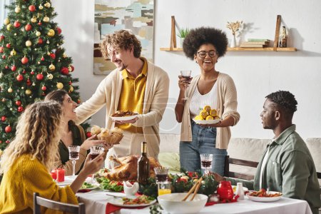 glückliche multiethnische Verwandte, die Spaß daran haben, Weihnachten zu feiern, lächeln und Teller mit Essen halten