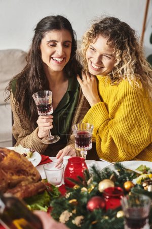 alegre lgbt pareja abrazándose unos a otros y sonriendo sinceramente, sosteniendo copas de vino, Navidad