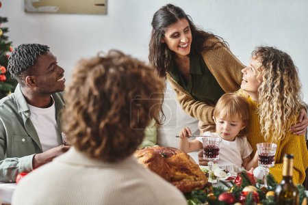 glückliche multiethnische Familienmitglieder, die am festlichen Tisch sitzen und viel Spaß haben, mit Tochter in den Händen