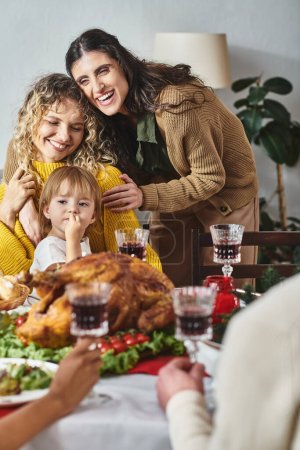 freudiges lgbt-Paar mit Tochter in den Händen, die fröhlich lächelt, während sie am Weihnachtstisch sitzt
