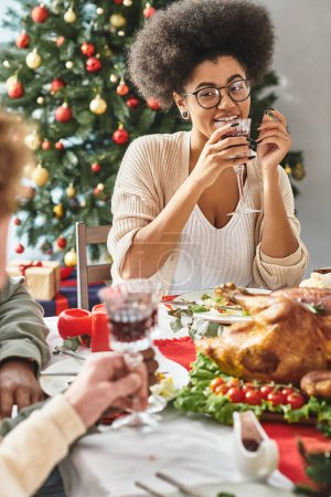 fröhliche afrikanisch-amerikanische Frau im Gespräch mit ihren Verwandten beim Weintrinken vor Weihnachtsbaumkulisse