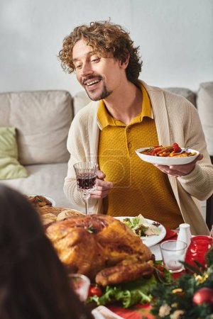 joven hombre alegre sentado en la mesa de vacaciones con su familia y disfrutando de la comida festiva, Navidad