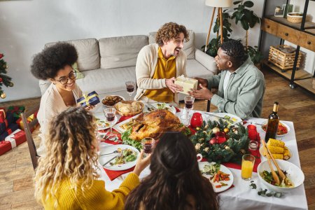 multiethnic family sitting at festive table with turkey and wine and exchanging gifts on Christmas