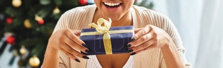Téléchargez les photos : Vue recadrée de heureuse femme afro-américaine tenant son cadeau de Noël et souriant sincèrement - en image libre de droit