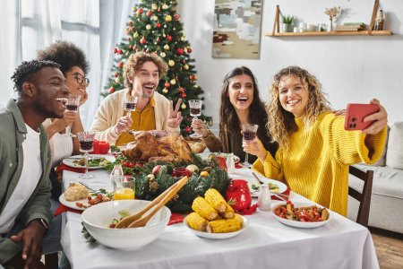 gran familia multirracial tomando selfie alegre sentado en la mesa festiva con árbol de Navidad en el telón de fondo