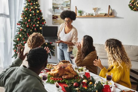 Multikulturelle Familie versammelte sich zum festlichen Mittagessen und winkte fröhlich lächelnd in die Laptop-Kamera, Weihnachten