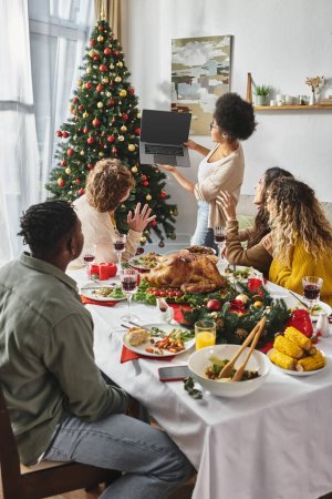Multikulturelle Familie beim festlichen Mittagessen und lächelnd in die Laptop-Kamera, Weihnachten