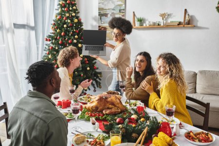 familia multiétnica hablando activamente y sonriendo a la cámara portátil en la mesa de vacaciones, Navidad