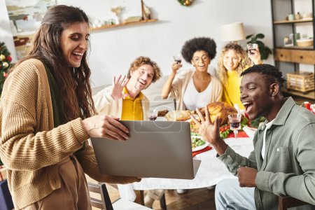 multiracial jolly relatives sitting at Christmas feast cheering and smiling at laptop camera