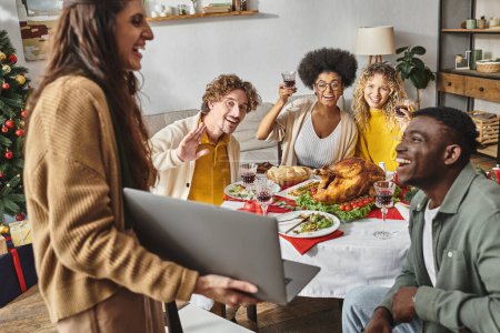 multiethnisch glückliche Familie, die beim Urlaubsessen mit Wein jubelt und in die Laptop-Kamera lächelt